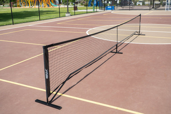 Portable low black net across an outdoor multi-use court, with playground and fencing visible in the background.